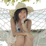 A person wearing a sunhat sits on a hammock by the beach, smiling with one hand resting on their chin.