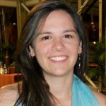 A woman with long brown hair and a light blue sleeveless top smiles at the camera in an indoor setting with plants and glass windows in the background.