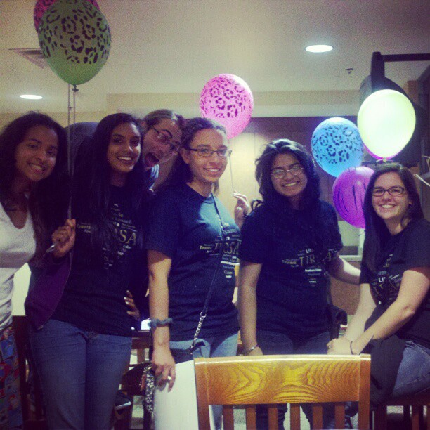 Six women smiling, standing and sitting indoors, holding colorful balloons, with chairs and tables visible in the background.