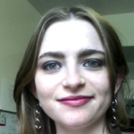 A woman with long brown hair and earrings smiles at the camera indoors, with a whiteboard and a window in the background.