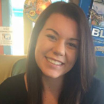 A young woman with straight brown hair and a black top smiles while sitting indoors, with colorful decorations visible in the background.