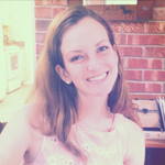 A woman with long brown hair smiles at the camera while sitting indoors in front of a brick wall.