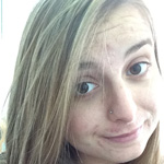 A person with long blonde hair and a small nose piercing looks at the camera with a slight smile.