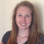 A young woman with long, straight, light brown hair smiles at the camera while standing against a plain, light-colored wall.