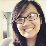 A person with long dark hair and glasses is smiling at the camera in a brightly lit room.