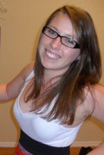 A young woman with straight brown hair and glasses is smiling, wearing a white top and black waistband, standing indoors against a beige wall.