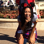 Woman wearing Minnie Mouse ears and a birthday button smiles and poses outdoors in a theme park, with other visitors and flowers in the background.