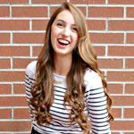 A woman with long, wavy brown hair wearing a white and black striped shirt smiles while standing in front of a brick wall.