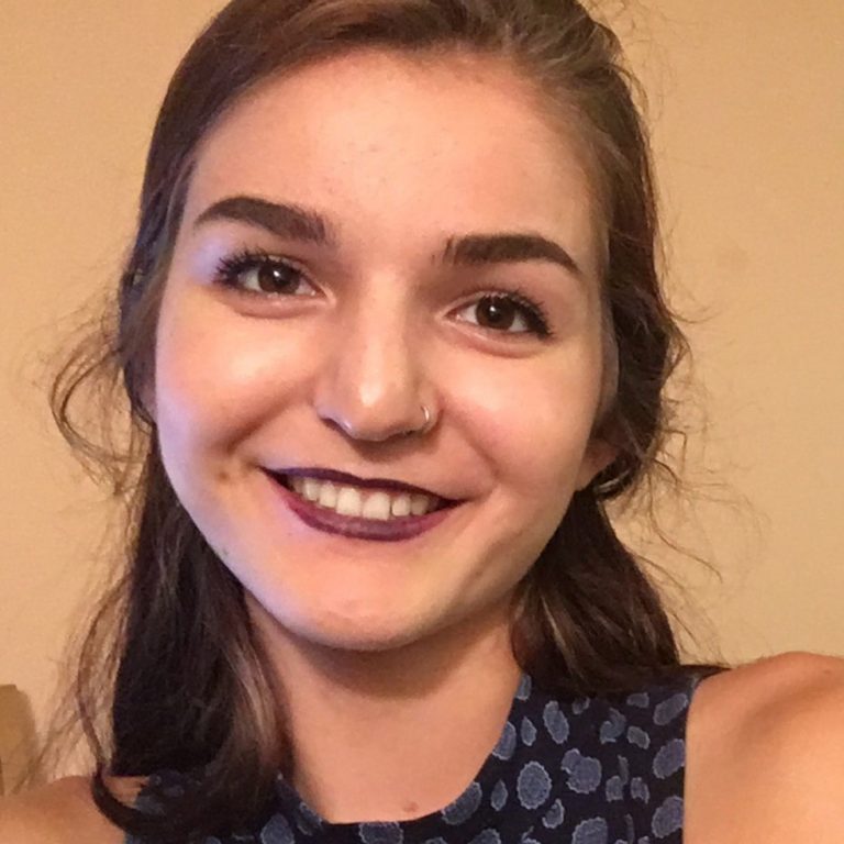 Young woman with long brown hair, wearing a blue patterned top, smiles at the camera. She has a nose ring and dark lipstick. The background is plain beige.