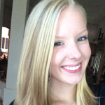 A young woman with straight blonde hair smiles at the camera indoors.