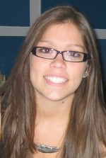 A woman with long brown hair and glasses smiles at the camera. She is wearing a necklace and is positioned in front of a blue and white background.