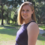 A woman with shoulder-length blonde hair, wearing a sleeveless dark top, stands outdoors on a sunny day with trees and grass in the background.
