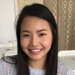 A woman with long dark hair smiles at the camera while sitting indoors, with patterned curtains in the background.