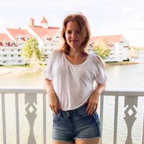 A person in a white shirt and denim shorts stands on a balcony overlooking water and white buildings with red roofs.