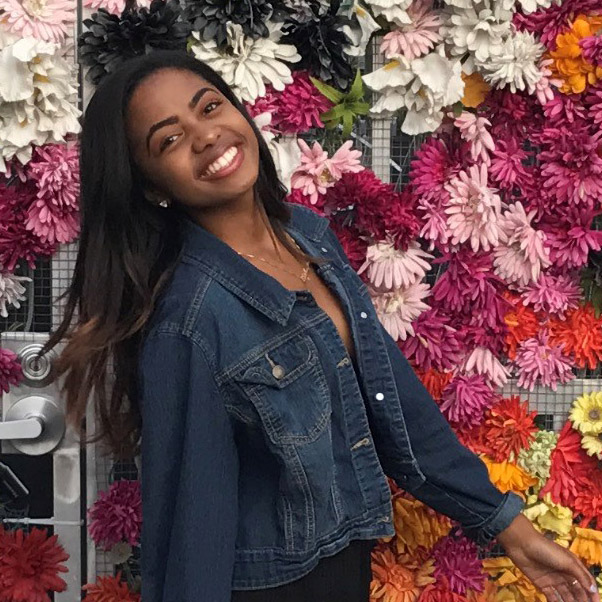 A woman wearing a denim jacket smiles and poses in front of a colorful wall decorated with artificial flowers.