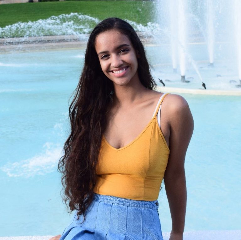 Woman with long dark hair wearing a yellow top and blue skirt sits in front of a fountain, smiling at the camera.