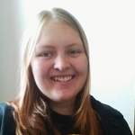 A person with long, straight, light brown hair is smiling at the camera while wearing a dark shirt, against a light background.