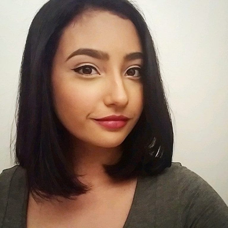 A woman with straight, shoulder-length dark hair and defined eyebrows looks at the camera, wearing a grey top and subtle makeup, against a plain background.