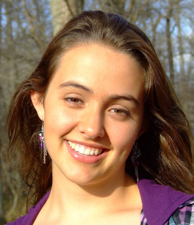 A young woman with long brown hair smiles outdoors, wearing dangle earrings and a purple top. Trees are visible in the blurred background.
