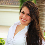 A woman with long dark hair, wearing a white shirt, smiles while standing outdoors in front of trees and a building.