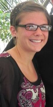 A person with short hair and glasses smiles at the camera, wearing a black and pink patterned top.