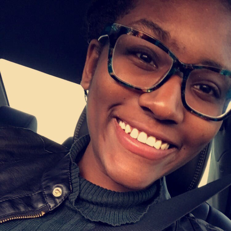 A person wearing glasses and a dark jacket smiles while sitting in a car, with a seatbelt visible across their chest.