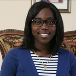 A woman with shoulder-length dark hair and glasses sits on a patterned chair, wearing a blue cardigan over a striped shirt.