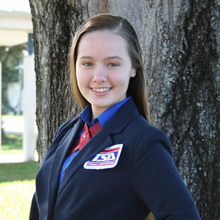 A young woman wearing a blazer with a TSA badge stands in front of a tree, smiling at the camera.