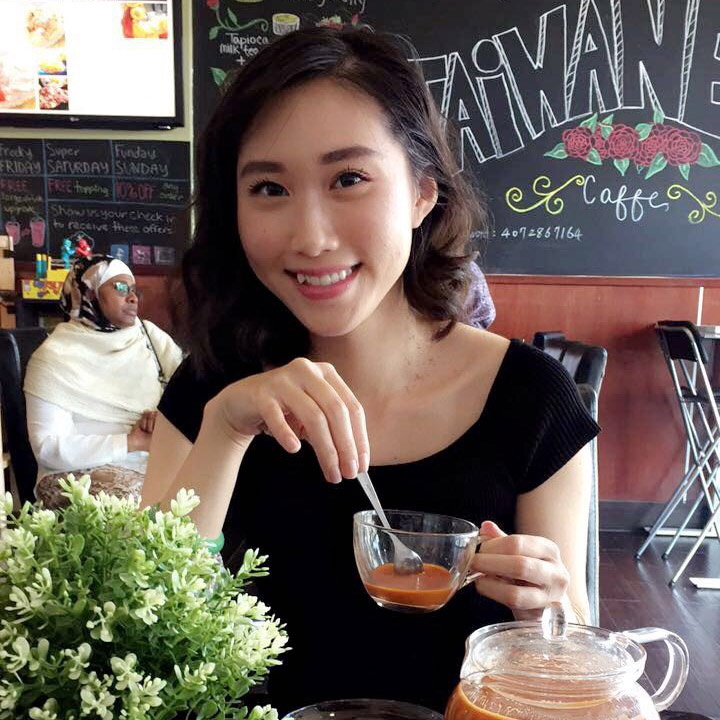 A woman sits at a table in a café, smiling at the camera while holding a glass teacup with tea. A teapot and potted plant are on the table. Café décor is visible in the background.