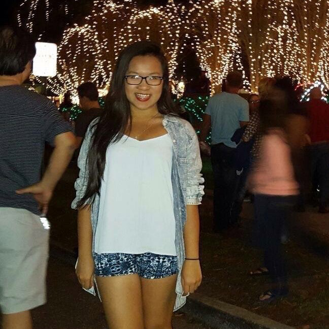 A young woman in glasses, a white top, and patterned shorts stands and smiles at night in front of people and trees decorated with string lights.