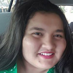 A person with long dark hair and a green top is smiling while sitting in a car.