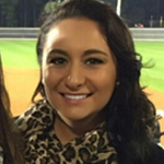 A woman with long dark hair, wearing a leopard print scarf, smiles outdoors with a blurry field and fence in the background.
