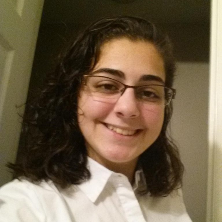A person with shoulder-length dark hair, glasses, and a white collared shirt smiles while standing indoors near a doorway.