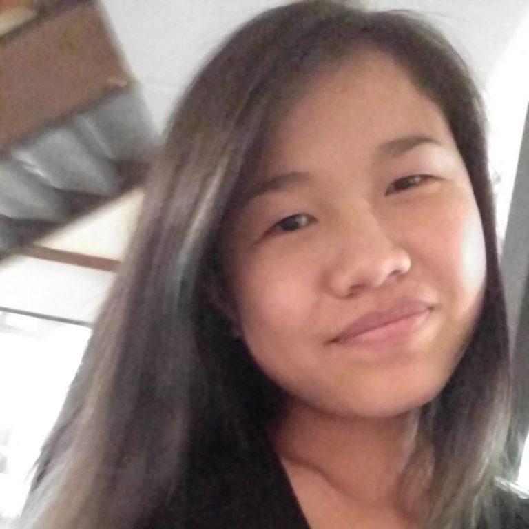 A person with long, straight dark hair is smiling slightly and looking at the camera indoors, with blurred background elements visible.