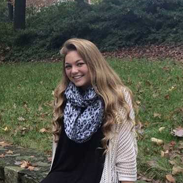 A young woman with long, wavy blonde hair sits outdoors on a stone ledge, smiling, wearing a black top, light cardigan, and a patterned scarf, with grass and fallen leaves in the background.
