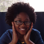 A person with curly hair and glasses smiles, resting their face in their hands, sitting at a table with a dark background.