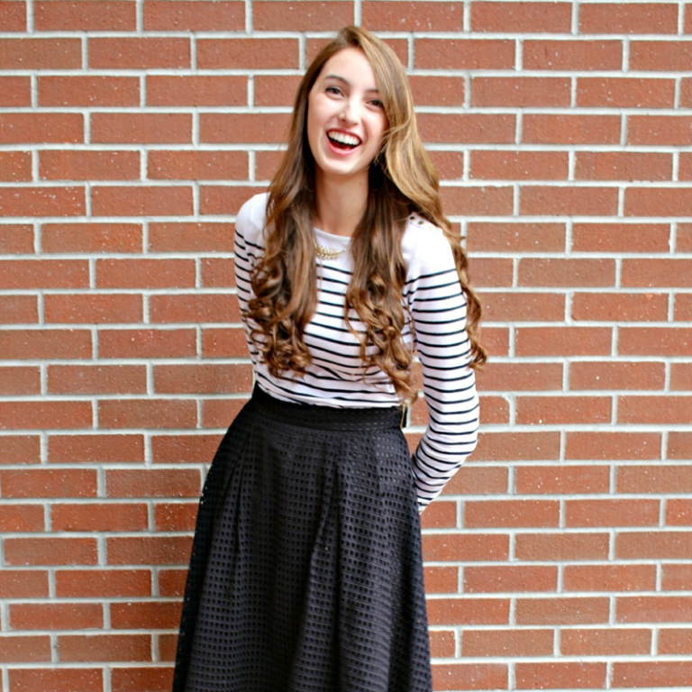 A young woman with long brown hair wearing a striped long-sleeve shirt and black skirt stands smiling in front of a brick wall.