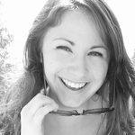 A woman smiles at the camera while holding sunglasses near her face in an outdoor setting. The photo is black and white.