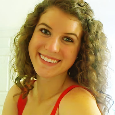 A woman with curly brown hair and a red top smiles at the camera against a light background.