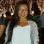 A woman with long dark hair and glasses smiles at the camera, standing outdoors at night with festive lights in the background.