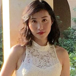 A woman with shoulder-length dark hair wearing a sleeveless white lace top stands outdoors with greenery and an archway in the background.