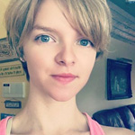 A person with short blonde hair and blue eyes is indoors, wearing a pink top, with wall decor and furniture visible in the background.