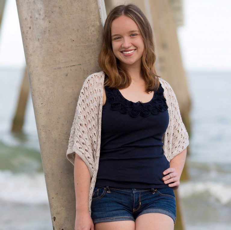 A young woman with brown hair, wearing a navy blue top, shorts, and a light knit cardigan, stands by a pier on the beach, smiling at the camera.