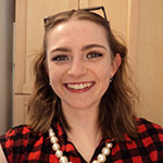 A young woman with wavy brown hair, wearing a red and black plaid shirt, a pearl necklace, and glasses on her head, smiles at the camera indoors.