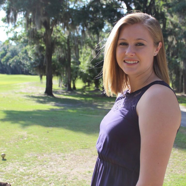 Woman with blonde hair in a sleeveless navy dress stands outdoors on a grassy field with trees in the background, smiling at the camera.