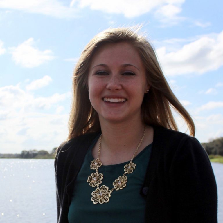 A woman with long hair smiles outdoors by a body of water, wearing a dark top, a black cardigan, and a gold floral necklace. The sky is bright and partly cloudy.
