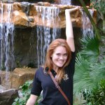A woman with red hair stands in front of a rocky waterfall, smiling and raising one arm above her head. She is wearing a black short-sleeve shirt and has a crossbody bag.