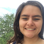 A woman with long brown hair smiles at the camera outdoors, with green trees and a partly cloudy sky in the background.