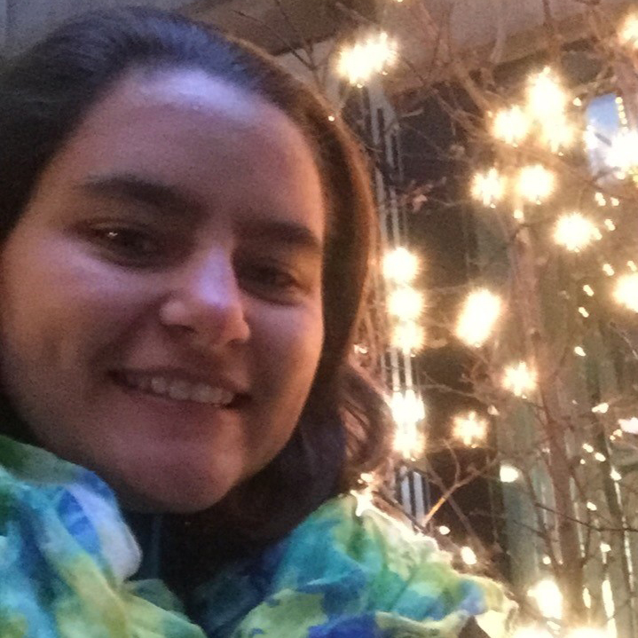 Person smiling at the camera in front of a tree decorated with glowing string lights at night.