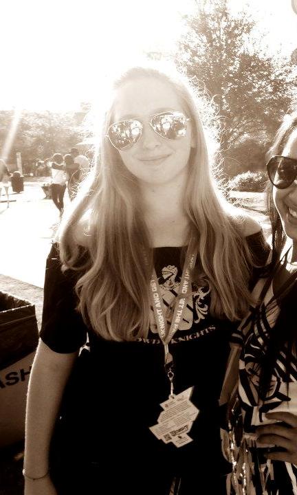 A young woman with long hair wearing sunglasses, a lanyard, and a dark shirt stands outdoors, smiling at the camera. Another person is partially visible beside her.